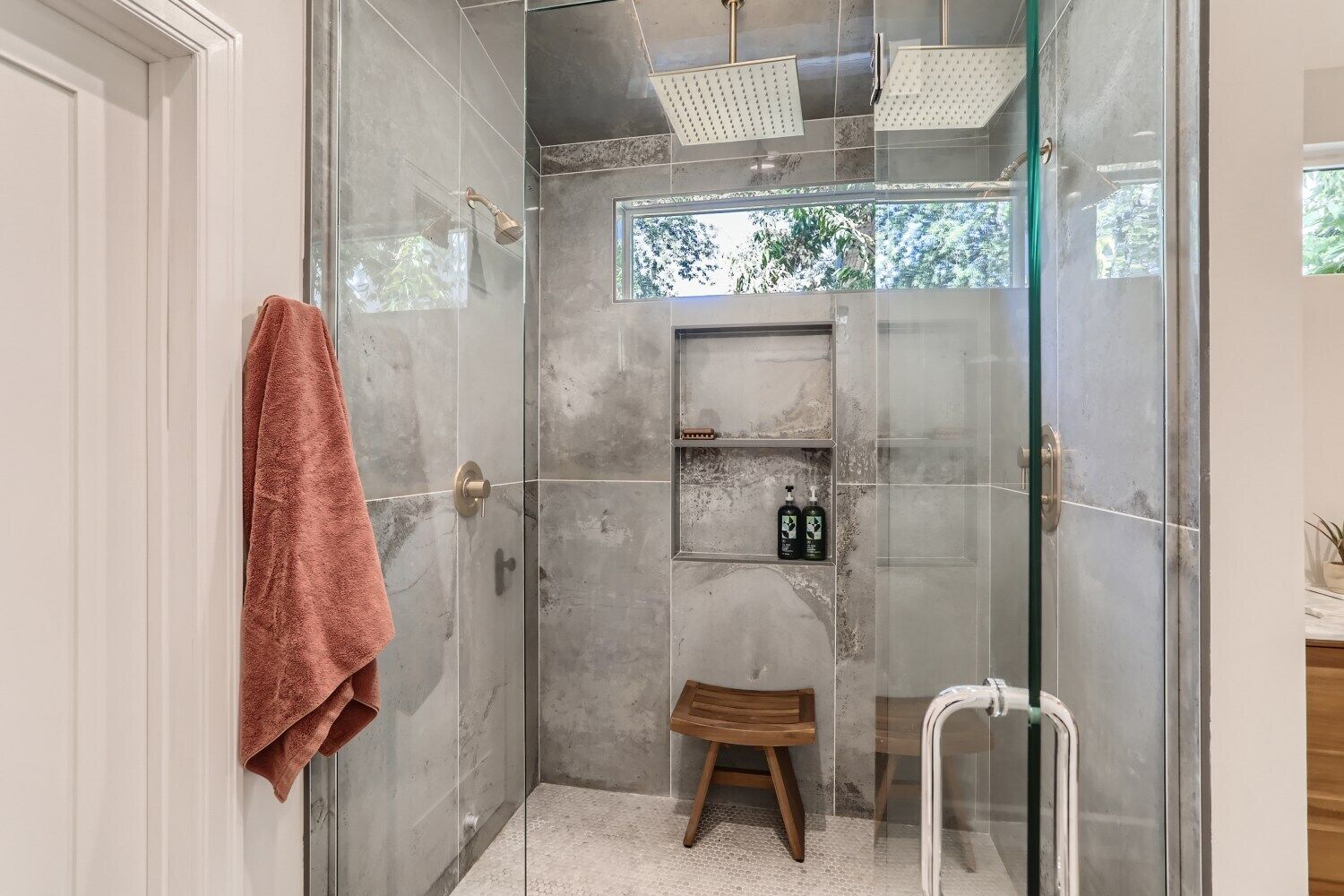 Une douche fermée par des parois de verre, avec un tabouret en bois, une serviette et une étagère contenant des articles de toilette.