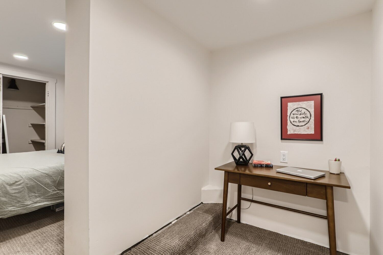 Un dormitorio pequeño y ordenado con cama, escritorio con lámpara y una cita inspiradora enmarcada en la pared.