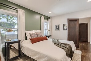 Un dormitorio con cama, mesitas de noche, una ventana con cortinas, una puerta y cuadros en la pared.
