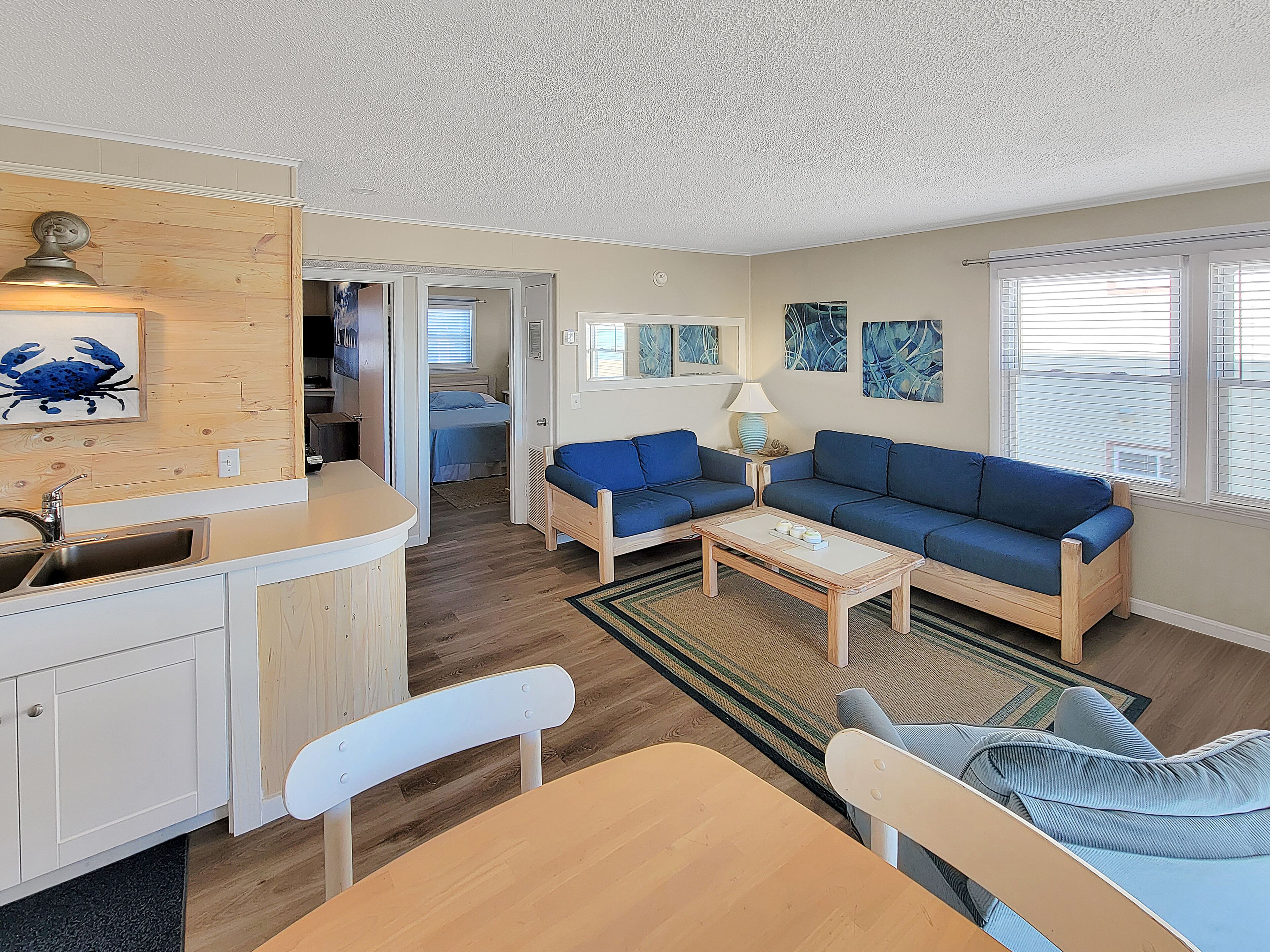 Un espacio habitable compacto con kitchenette, un sofá azul, una mesa de comedor y un pequeño recibidor que da a un dormitorio.
