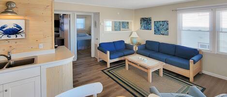 Un espacio habitable compacto con kitchenette, un sofá azul, una mesa de comedor y un pequeño recibidor que da a un dormitorio.