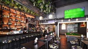 A bar area with wooden shelves stocked with bottles, a pool table, and a television displaying a sports game. - KOKO Hostel- Adults Only  (Krabi)