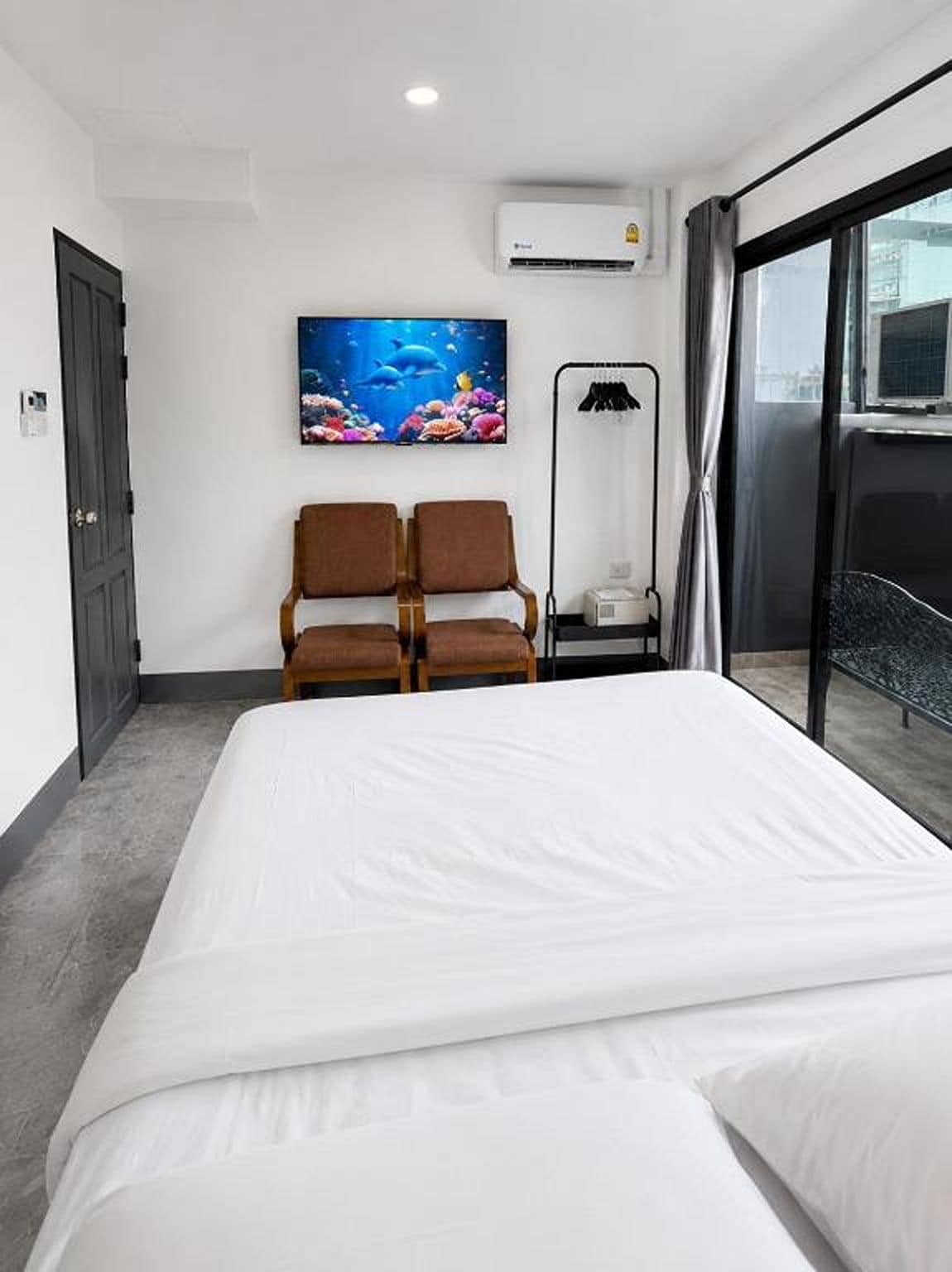 A hotel room with a bed, two chairs, a painting, an air conditioner, and a view of the outside.