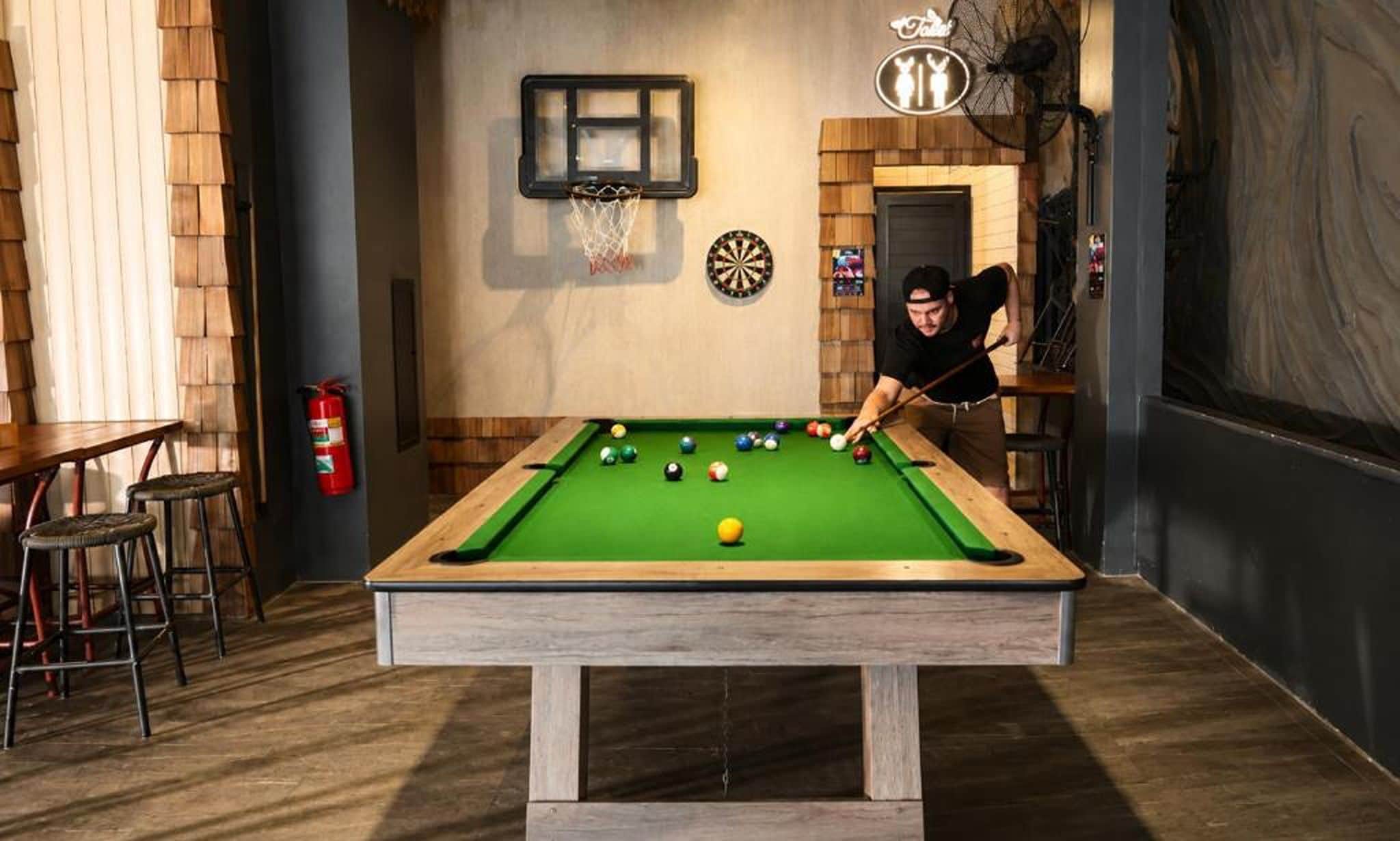 A pool table with balls, a dartboard, and a basketball hoop in a room with wooden walls and a fire extinguisher.