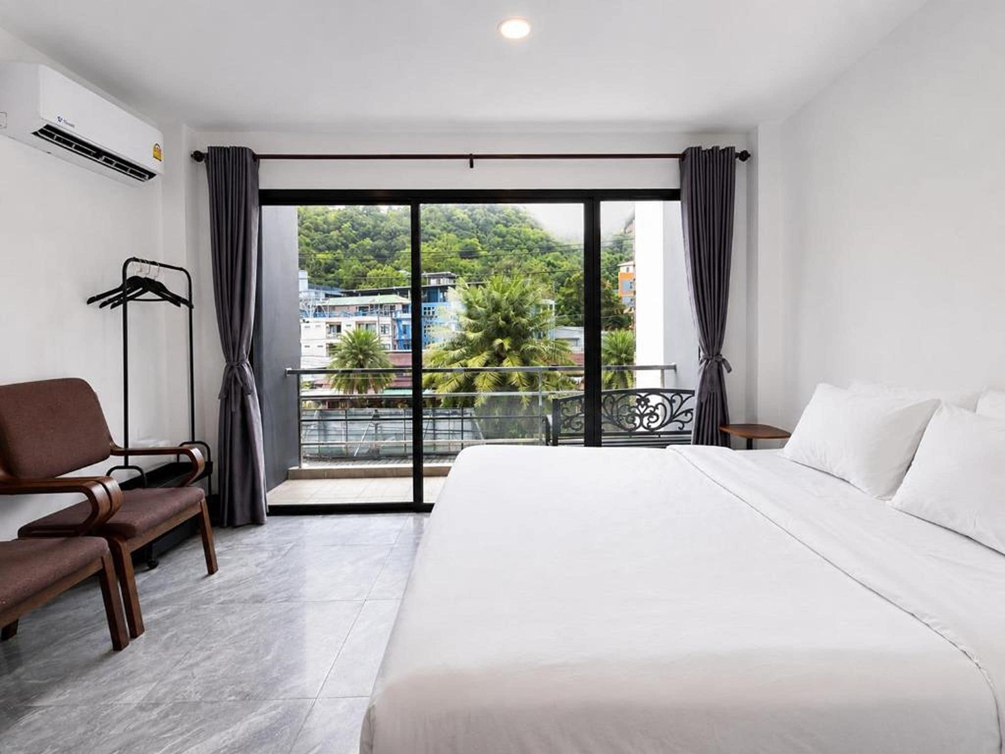 Ein Hotelzimmer mit einem großen Bett, einer Sitzecke mit einem Sessel, einem kleinen Tisch und einem Balkon mit Blick auf Gebäude und Grünflächen.