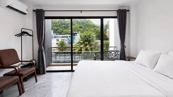 Ein Hotelzimmer mit einem großen Bett, einer Sitzecke mit einem Sessel, einem kleinen Tisch und einem Balkon mit Blick auf Gebäude und Grünflächen.