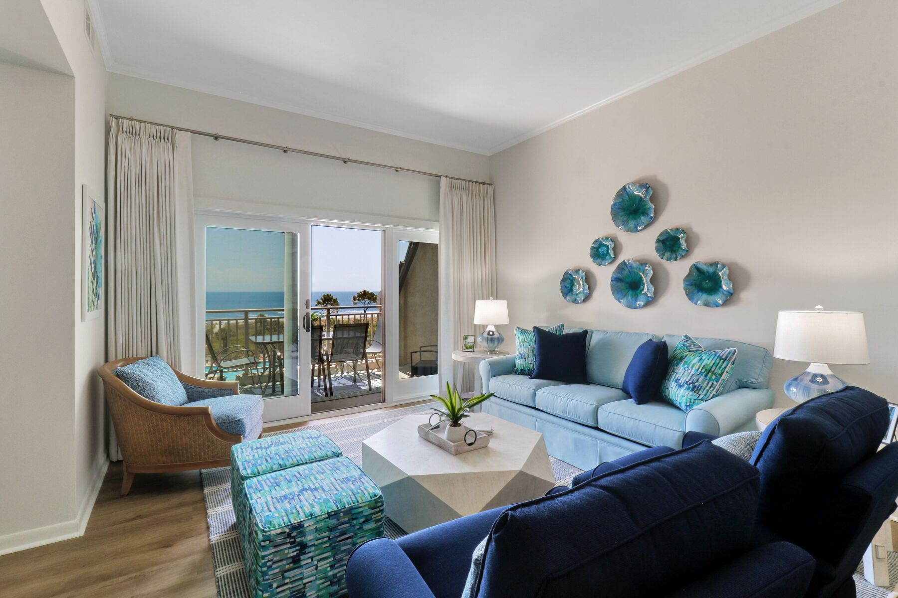 A living room with a light blue sofa, a wicker chair, a coffee table, and a view of the ocean through a sliding glass door.