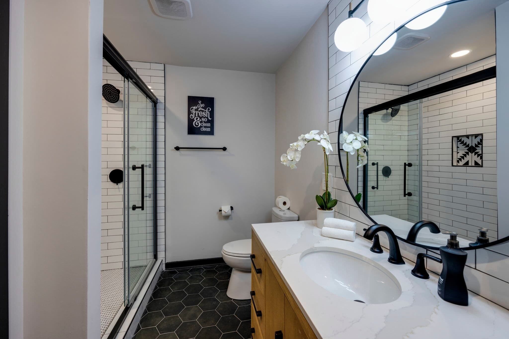 A modern bathroom with a glass shower enclosure, a white toilet, a large mirror with lights, and a white sink with black faucets.