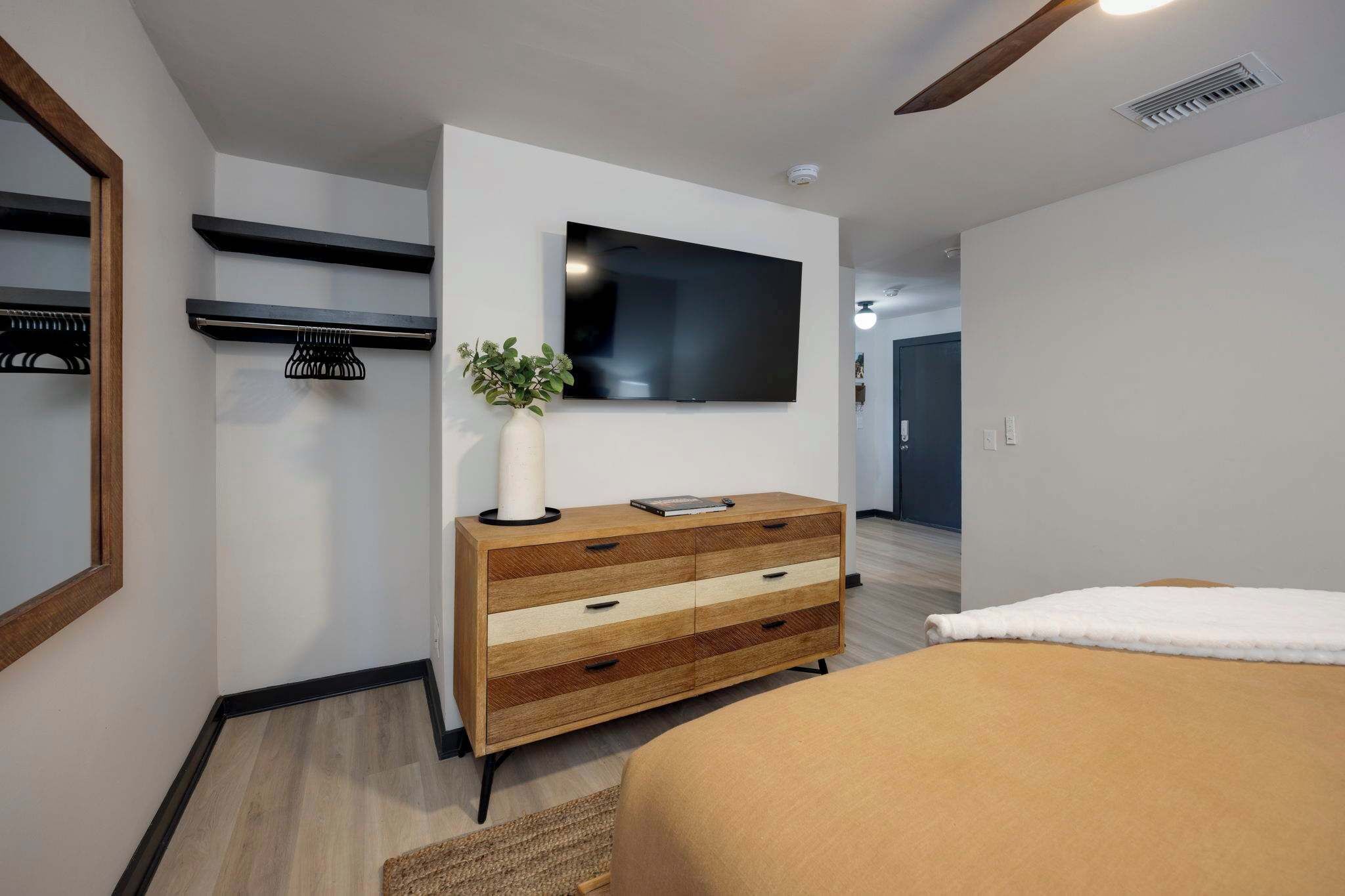 Een moderne hotelkamer met een flatscreen televisie aan de muur, een houten dressoir met lades, een nachtkastje en een groot bed met een witte deken.