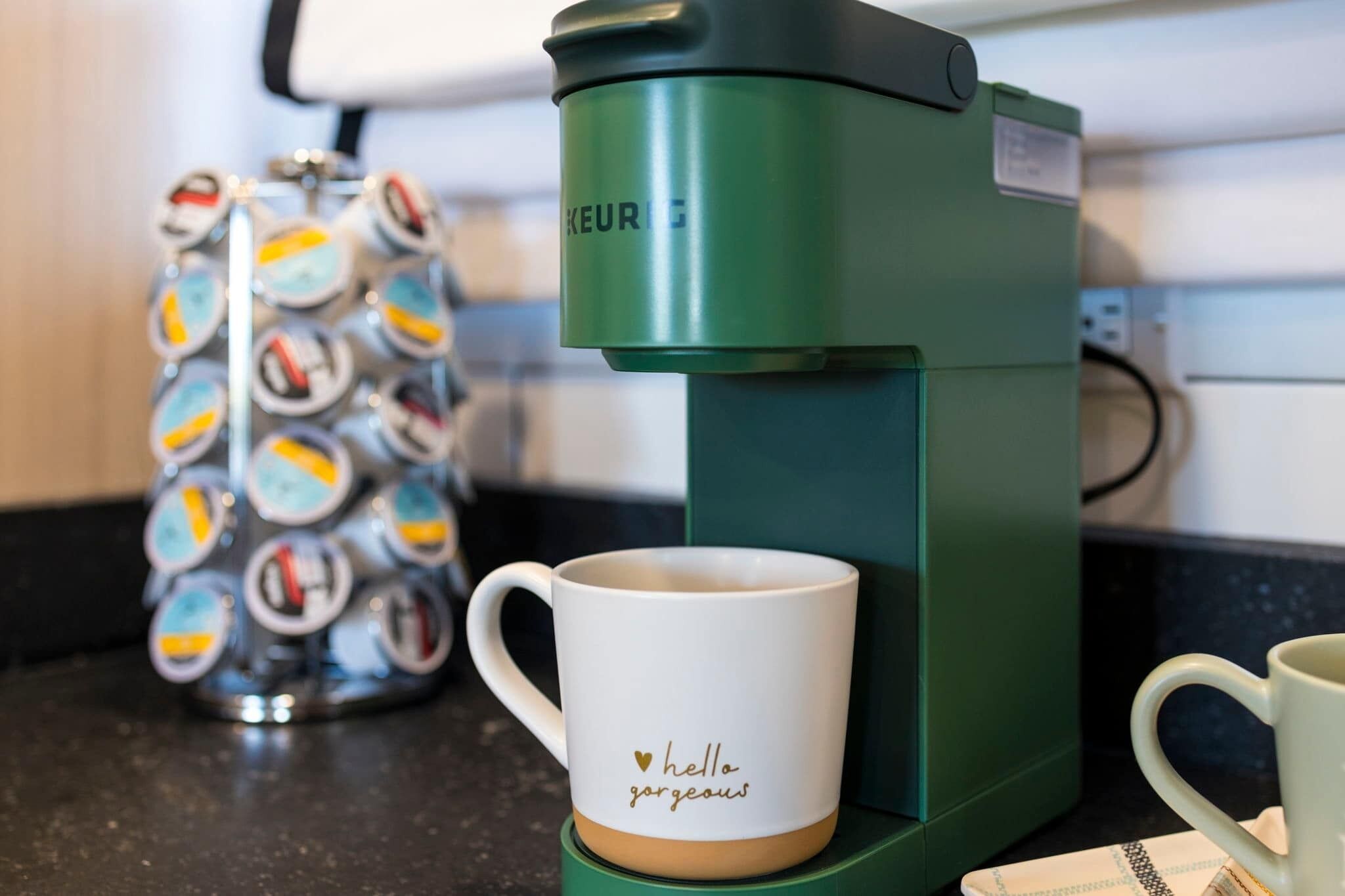 A coffee maker, a mug with "hello gorgeous" text, and a tin of coffee pods on a kitchen counter.