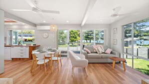 A modern kitchen with white cabinets, a dining area with a round table and chairs, a grey sofa with cushions, and large windows offering a view of the outdoors. - Sunset Sails Taylors Beach NSW (Taylors Beach)