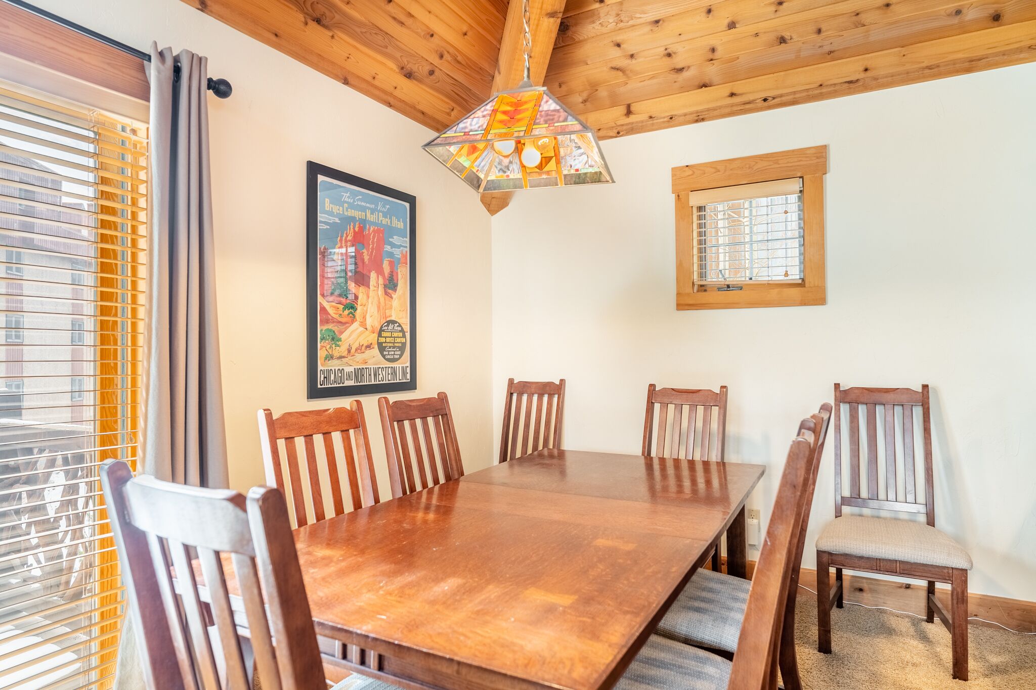 A dining area with a wooden table and chairs, a stained glass light fixture, a framed poster on the wall, and a window with blinds.