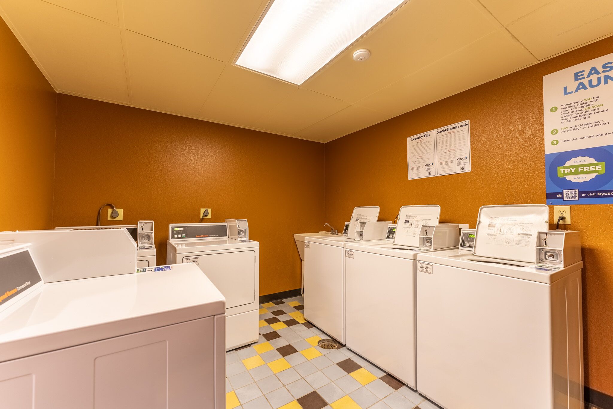 A laundry room with multiple washing machines and dryers, a tiled floor, and a sign advertising a new launch.