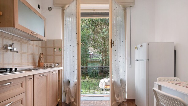 Une cuisine avec des meubles en bois, un plan de travail, un réfrigérateur et une vue sur un jardin, visible à travers une porte ouverte.