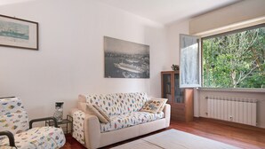 Un salon comprenant un canapé, des fauteuils, un tableau encadré, un meuble et une fenêtre donnant sur un paysage verdoyant.