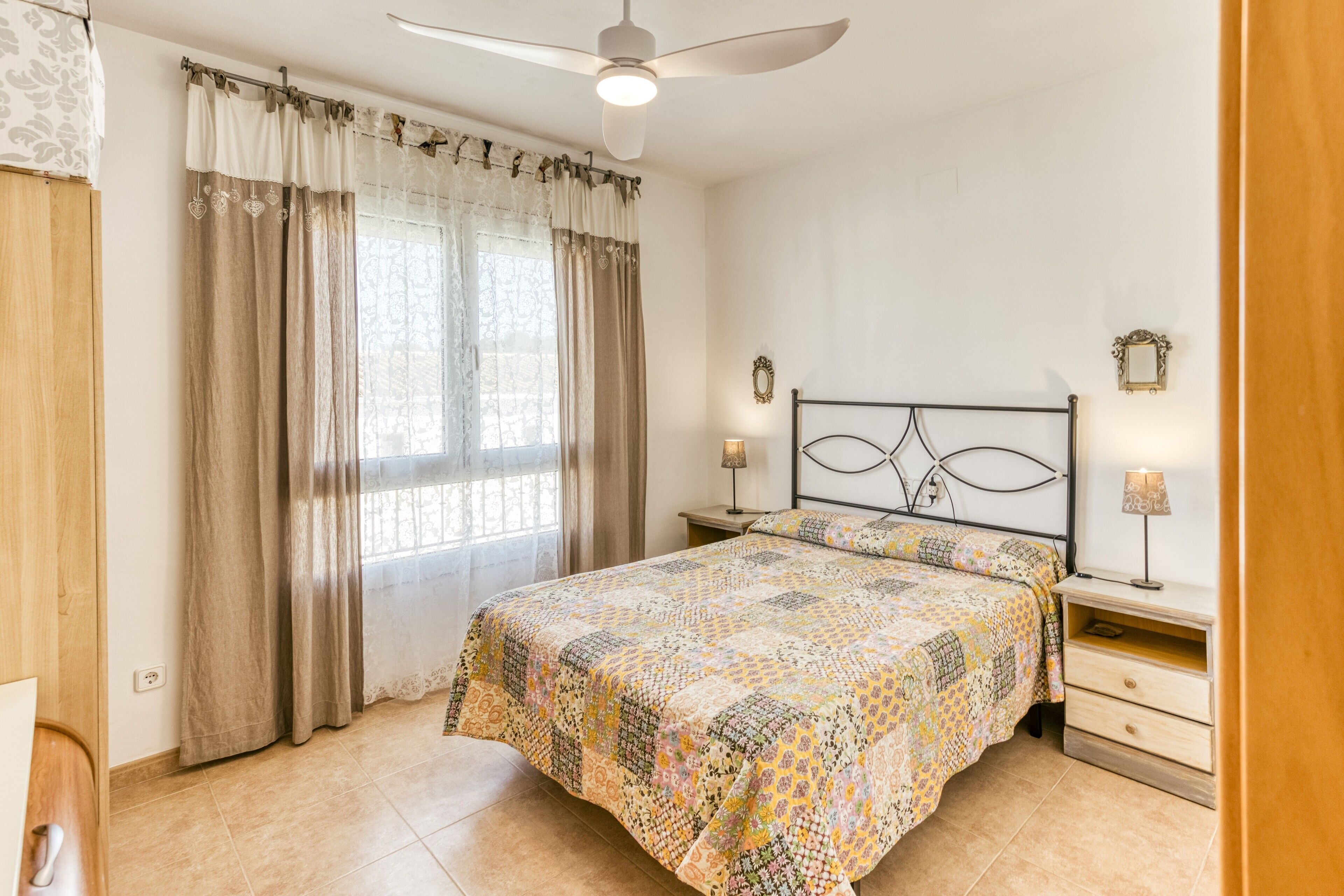 A bedroom with a bed, curtains, a ceiling fan, bedside lamps, and a nightstand.