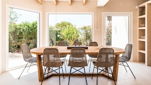 Un espace repas avec une table ronde et des chaises en osier, de grandes fenêtres et un plafond en bois.
