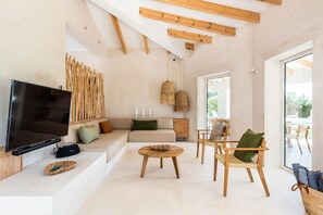 A modern living room with a flat-screen TV, wooden furniture, a sofa with cushions, and a large window.