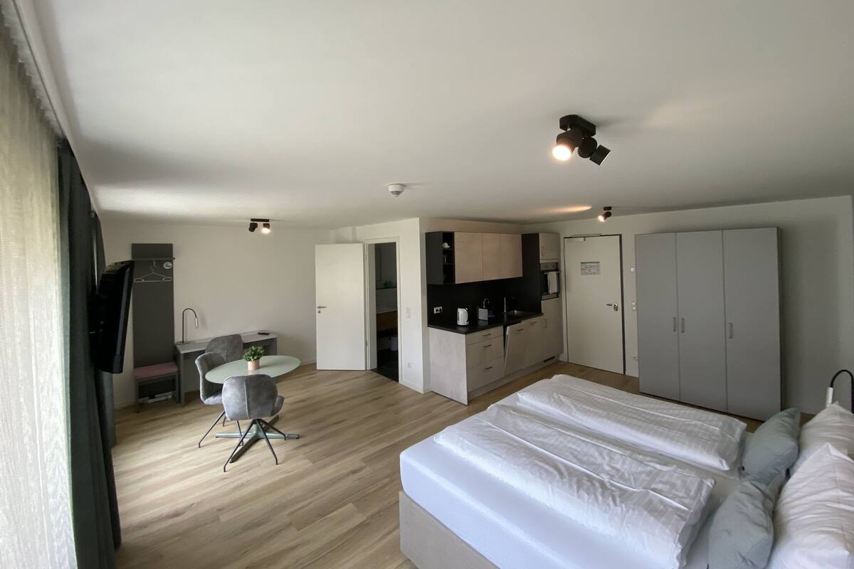 A modern hotel room with a large bed, a kitchenette, a seating area with a table and chairs, and a wardrobe.
