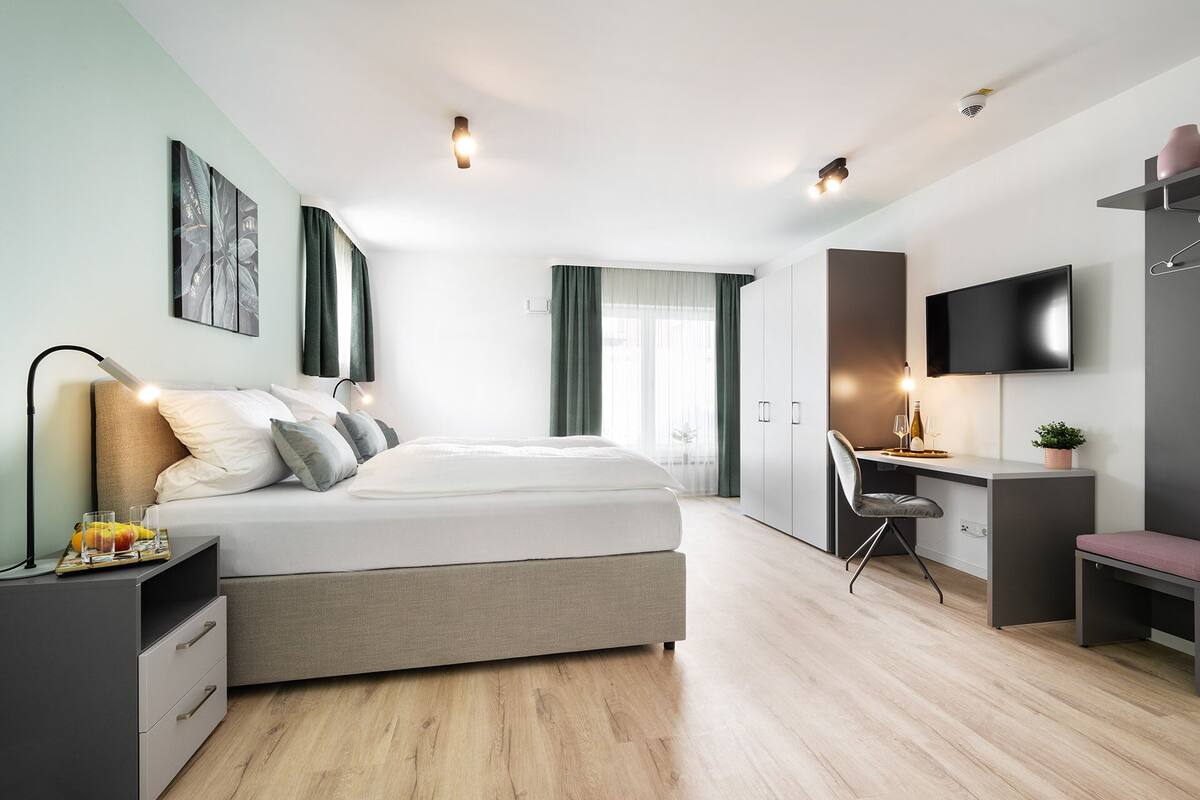 A modern bedroom with a bed, bedside tables, a desk with a chair, a TV, and a plant.