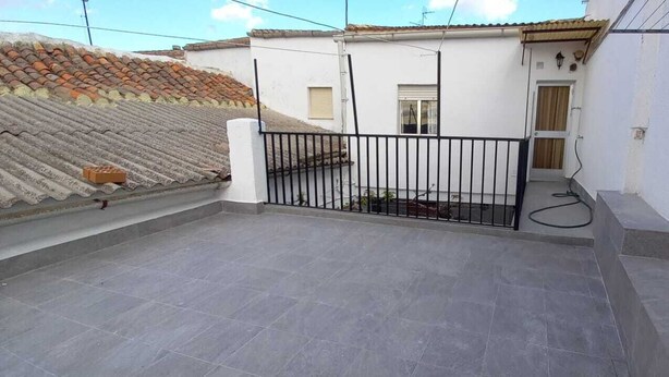 Una terraza en la azotea con suelo de baldosas, una reja metálica negra y un edificio blanco con una puerta y una ventana.