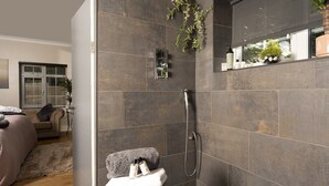 A modern bathroom with dark tiles, a shower, a towel rack, and a small shelf with toiletries. - Stay at-FL (Crawley)