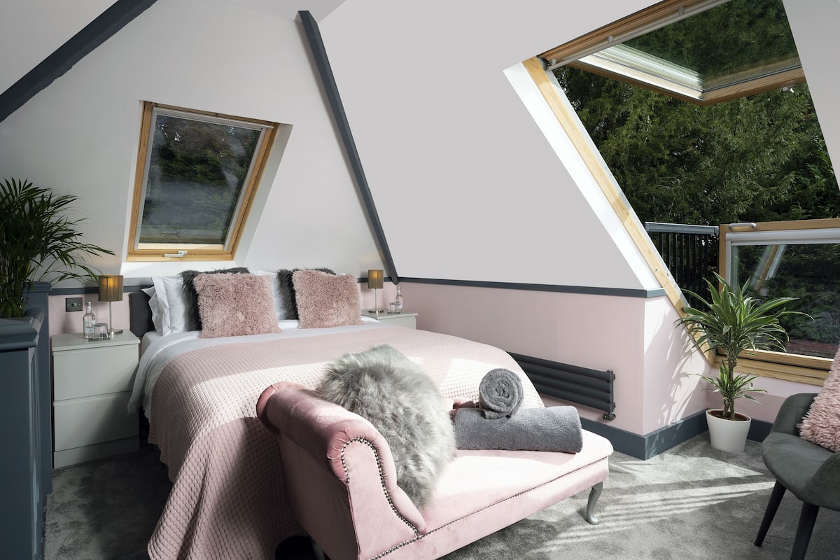 A bedroom with a sloped ceiling, a bed with pink bedding, a bedside table with a lamp, and a window with a view of trees.