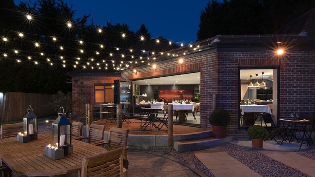 An outdoor dining area with string lights, wooden furniture, and a brick building with a glass door. - Stay at-FL (Crawley)