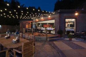 Un espacio de comedor al aire libre con luces colgantes, muebles de madera y un edificio de ladrillo con puerta de vidrio.