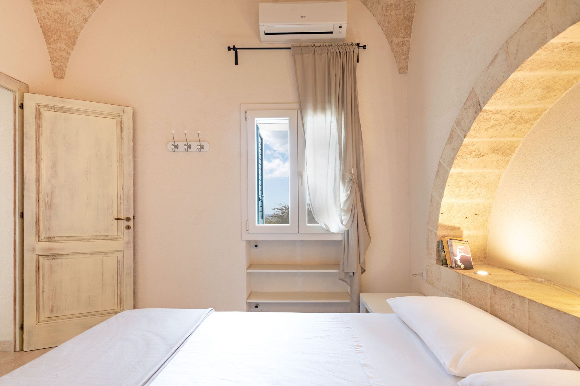 Une chambre avec un lit, une fenêtre avec des rideaux, un appareil de climatisation et un mur en pierre.