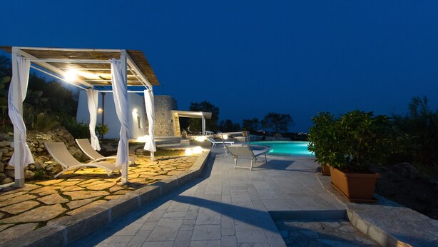 A stone-paved outdoor area with a pool, lounge chairs, and a pergola with lights.