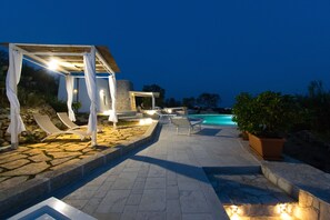 A stone-paved outdoor area with a pool, lounge chairs, and a pergola with lights. - VILLA Alessandra (Marina di Pescoluse)
