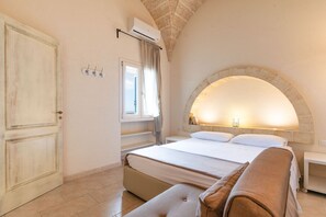 Un dormitorio con cama, un banco, una mesita de noche, una pared de piedra con arco, una ventana con cortinas y un equipo de aire acondicionado.
