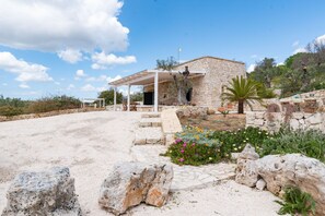 Una casa de piedra con un porche cubierto, rodeada por un jardín con diversas plantas y flores.