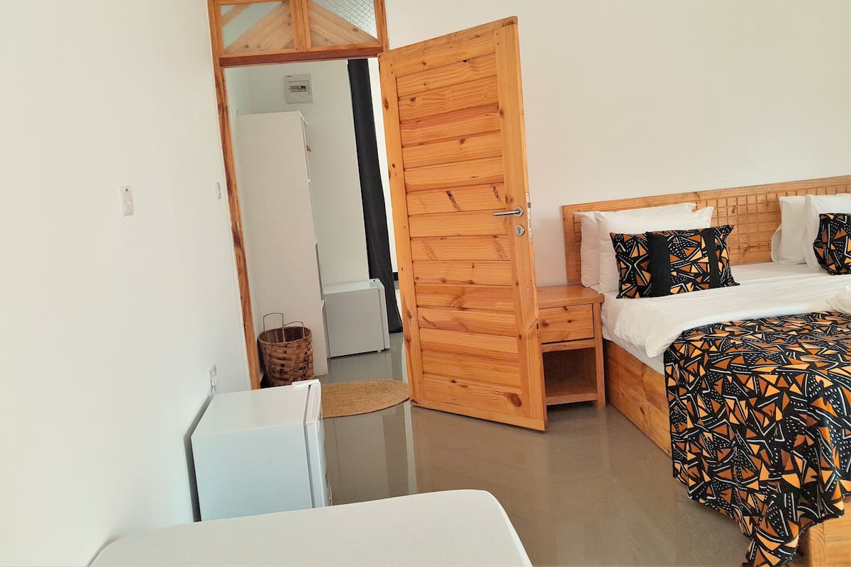 Un dormitorio con un armario de madera, una cama con ropa de cama estampada, una mesita pequeña blanca y un refrigerador blanco.