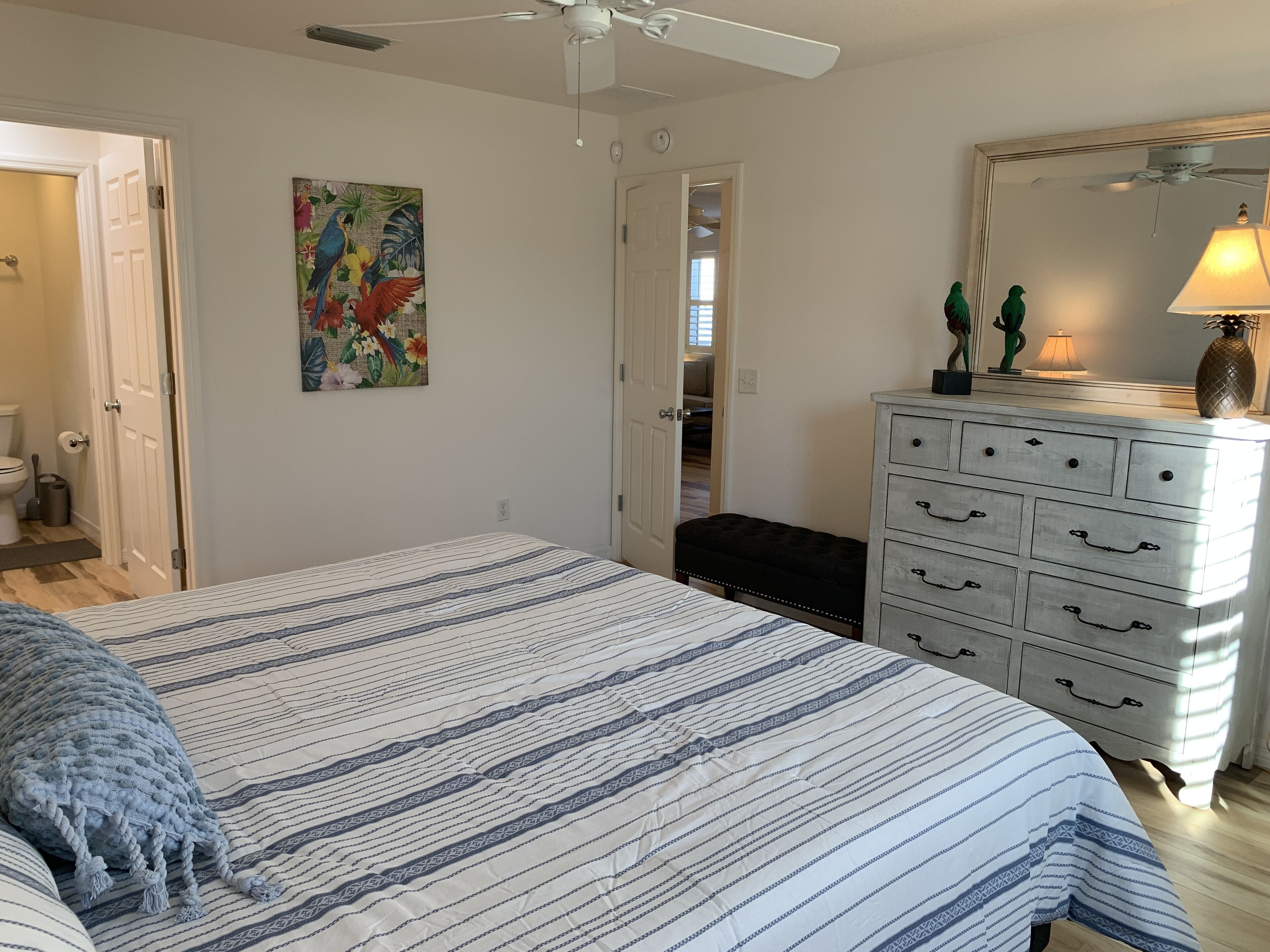 A bedroom with a bed, a dresser, a ceiling fan, a painting, and a door leading to another room.