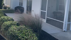 A single-story house with a sliding glass door, a covered patio area, and a small garden with bushes. - Beautiful 2/2 Villa w/4 Seater Gas Golf Cart, BBQ Grill, and WiFi!! (The Villages)