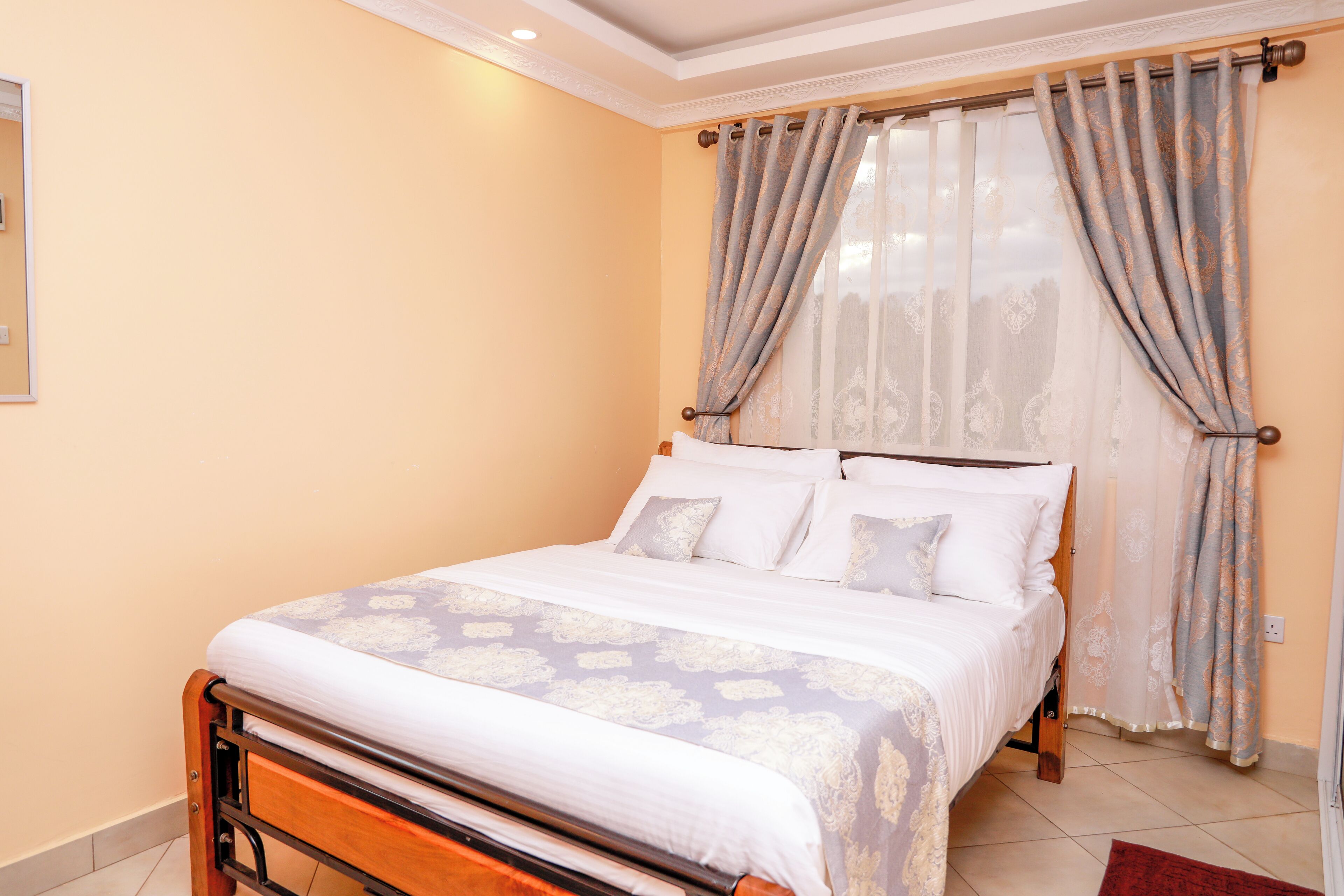 A neatly made bed with a patterned bedspread and pillows, a wooden headboard, and a window with sheer curtains.