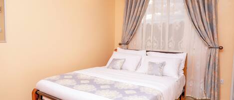A neatly made bed with a patterned bedspread and pillows, a wooden headboard, and a window with sheer curtains.