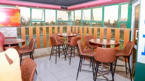 Een restaurant met meerdere tafels en stoelen, grote ramen met uitzicht op groen, en een roze plafond.