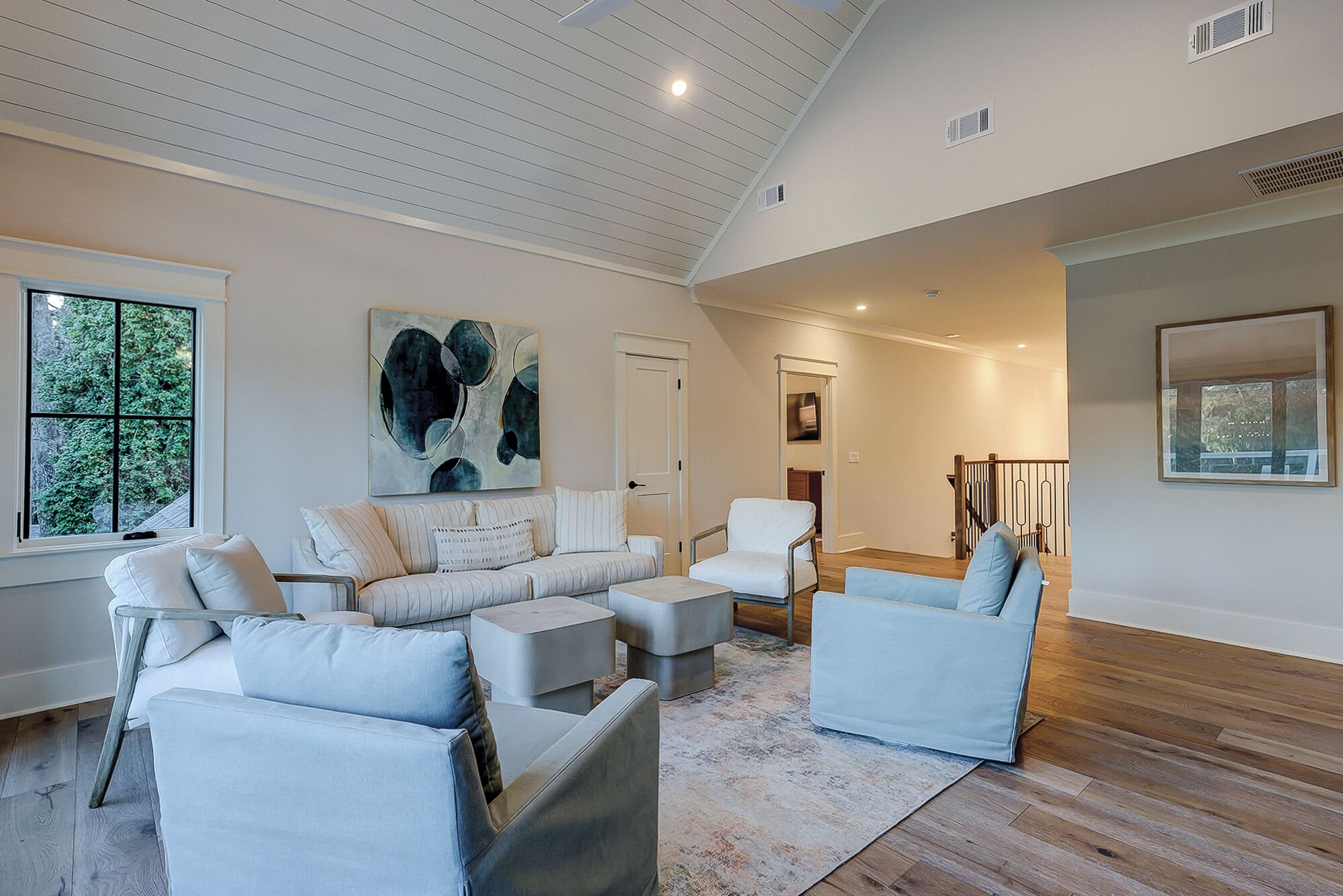 A spacious living room with a high ceiling, a large abstract painting, and a view of the outdoors through a window.