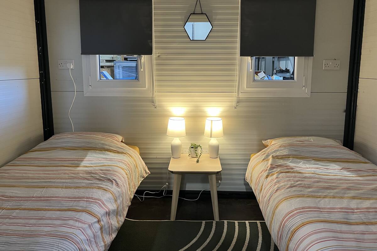 A room with two beds, each with striped bedding, a small table with two lamps, and a hexagonal light fixture above.