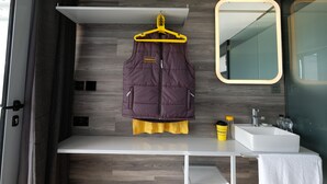 Une petite salle de bain avec un plancher en bois, un lavabo blanc, un miroir et un vêtement suspendu  à un cintre jaune.