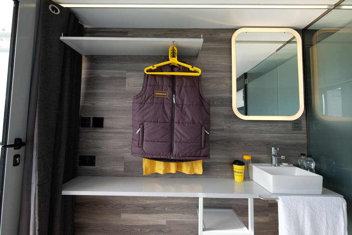 A compact bathroom with a wooden floor, a white sink, a mirror, and a hanging garment on a yellow hanger.