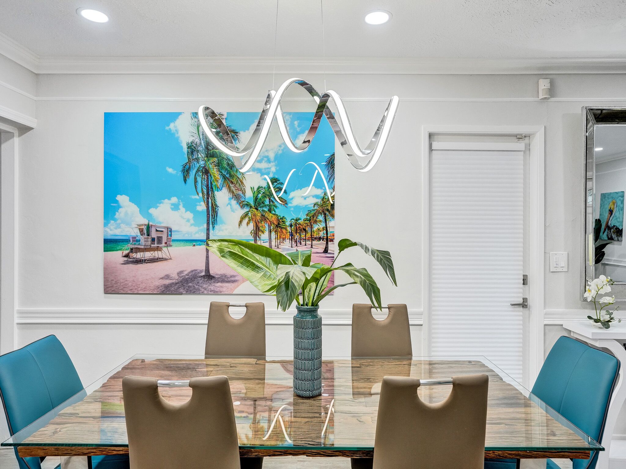 Un coin repas avec une table en verre, des chaises de couleur beige, un grand tableau représentant une scène de plage et un luminaire moderne.