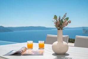 A table with a vase of flowers, two glasses of orange juice, and a magazine on a terrace overlooking a body of water.