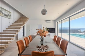 Un comedor moderno con una mesa larga, sillas naranjas, un jarrón con flores, una escalera y vistas a una piscina.