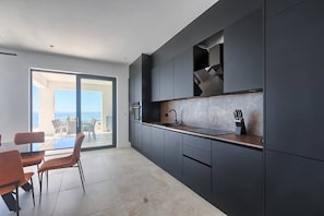A modern kitchen with dark cabinetry, a countertop, and a sink. There is a dining area with orange chairs and a table. - Villa Ultima with sea view, private pool&jacuzzi (Ripenda Kosi)