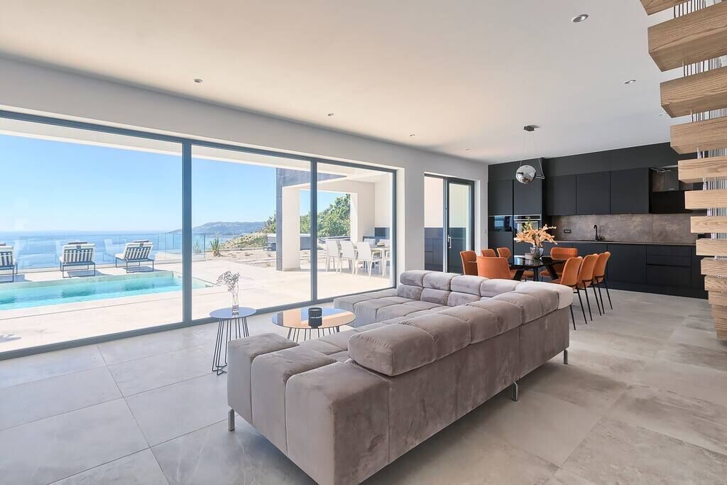 A modern living room with a large sofa, a dining table, and chairs, a kitchen area, and a view of the ocean.