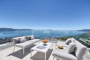 A balcony with a glass railing, two grey sofas, a white coffee table, and a view of the sea and hills. - Villa Ultima with sea view, private pool&jacuzzi (Ripenda Kosi)